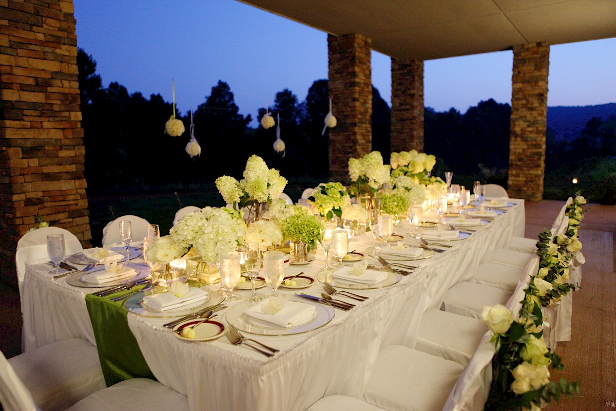 An elegantly set outdoor dining table with white flowers, candles, and white-covered chairs, against a backdrop of columns and evening sky.
