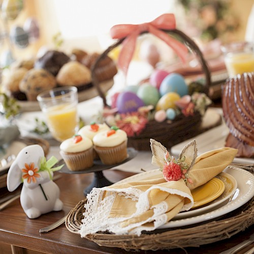 Festive table setting with Easter eggs, cupcakes, ham, and a ceramic bunny, surrounded by drinks and flowers.