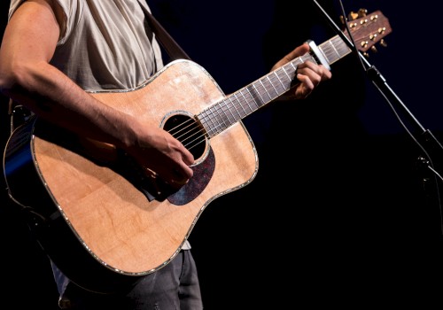 A person is playing an acoustic guitar on stage, holding a pick while wearing a casual shirt, with a microphone visible in the background.