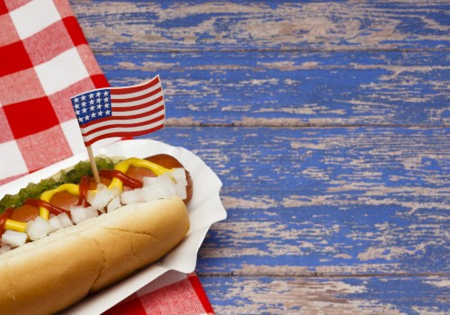 The image shows a hot dog with mustard, ketchup, onions, and relish in a paper tray, next to a small U.S. flag on a checkered tablecloth.