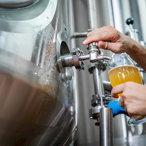 Someone filling a glass with beer from a large stainless steel brewing tank in a brewery setting.