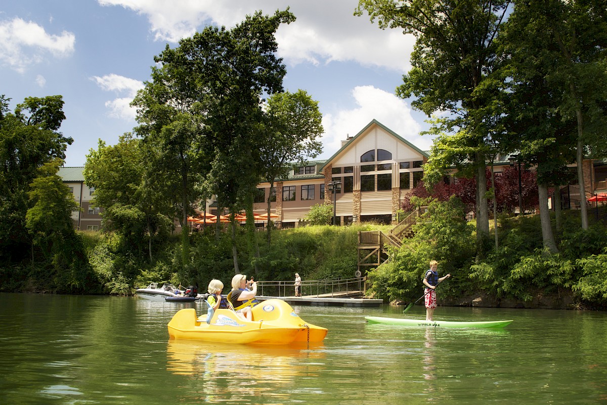 People enjoying a sunny day on the water with paddle boats, surrounded by greenery and a building in the background.
