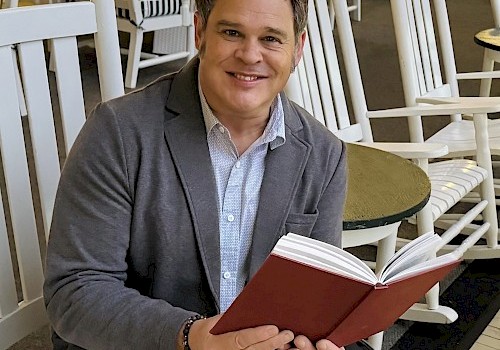 A person is sitting in a chair, smiling, while holding an open book in a setting with several similar chairs around.