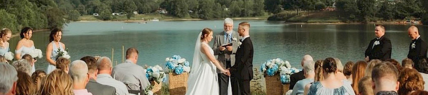 A wedding ceremony takes place outdoors by a lake, with guests seated and a scenic backdrop of hills and water, creating a serene setting.