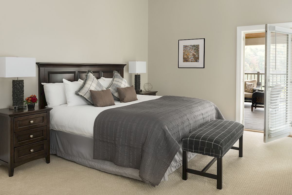 A bedroom with a large bed, bedside tables with lamps, a bench at the foot of the bed, and a door leading to a balcony or patio.